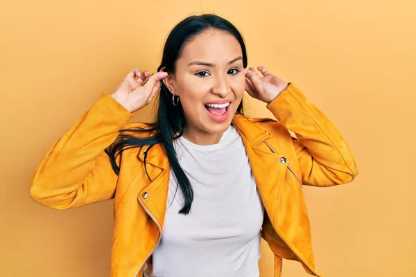 Beautiful hispanic woman with nose piercing wearing yellow leather jacket smiling pulling ears with fingers, funny gesture. audition problem