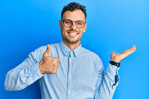 Latin asıllı genç adam günlük kıyafetler ve gözlük takıyor avuç içi gösteriyor ve baş parmaklarıyla iyi bir jest yapıyor, mutlu ve neşeli gülümsüyor. 