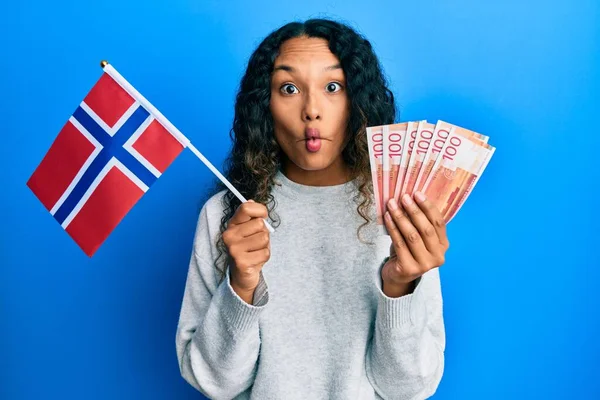 年轻的拉丁女子举着诺威国旗和挪威克朗钞票 用嘴和眼神做鱼脸 疯狂而滑稽 — 图库照片