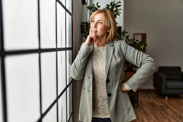 Middle age businesswoman smiling happy leaning on the window at the office.