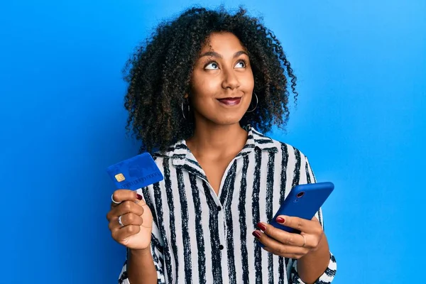 Beautiful african american woman with afro hair holding smartphone and credit card smiling looking to the side and staring away thinking.