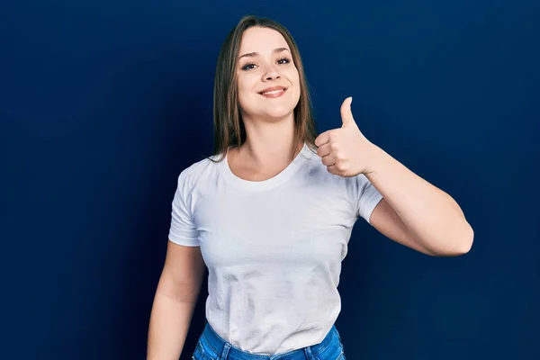若いヒスパニック系の女の子は手で幸せな親指アップジェスチャーを行うカジュアルな白いTシャツを着て 成功を示すカメラを見ると — ストック写真
