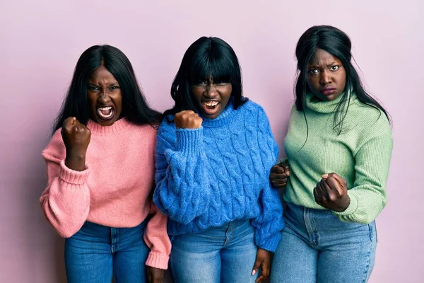 Three young african american friends wearing wool winter sweater angry and mad raising fist frustrated and furious while shouting with anger. rage and aggressive concept.