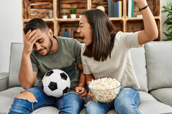 Young latin couple watching soccer match eating porpcorn at home.