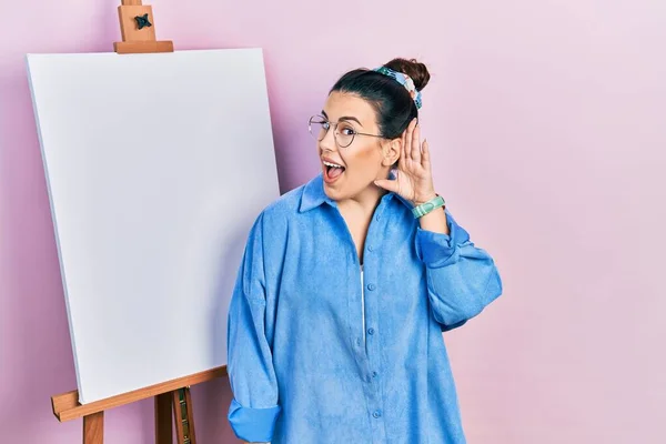画家のイーゼルのそばに立つ若いヒスパニック系の女性は 噂やゴシップに耳を傾け手で笑みを浮かべて立っている 聴覚障害の概念 — ストック写真