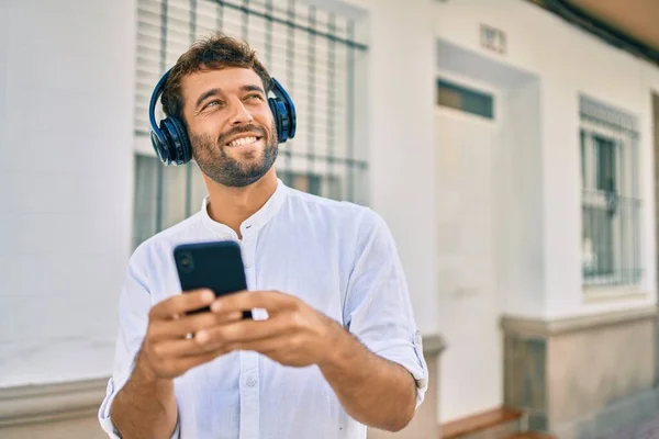 晴れた日に白いシャツを着て髭を生やしたハンサムな男は ヘッドフォンを着て音楽を聞いてスマートフォンを使って屋外で幸せな笑顔 — ストック写真