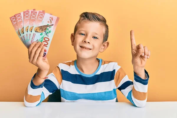 愛らしいですCaucasian子供保持100新しいニュージーランドドル紙幣上のテーブルの上に座っ笑顔でアイデアや質問を指差す指で幸せな顔 ナンバーワン — ストック写真