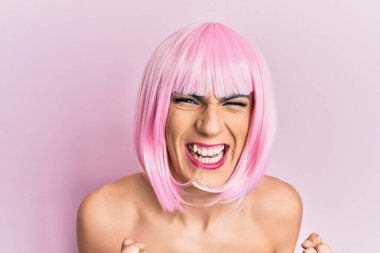 Pembe peruk takan genç adam mutlu ve heyecanlı bir şekilde kolları havada, gülümseyen ve başarı için çığlık atan kazanan bir jest yapıyor. kutlama konsepti. 