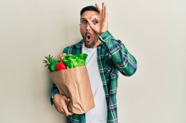 Sakallı yakışıklı bir adam, elinde torbayla market alışverişi yapıyor. Şaşırmış yüz ifadesi, parmaklarından bakan gözler. İnançsız ifade. 
