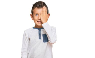 Sevimli beyaz çocuk günlük kıyafetler giyip bir gözünü eliyle kapatıyor, yüzünde kendine güvenen bir gülümseme ve şaşırtıcı bir duygu var.. 