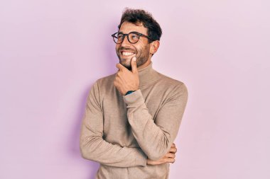 Balıkçı yaka kazak giyen ve eli çenesinde gözlük takan sakallı yakışıklı bir adam soru sormayı düşünüyor, düşünceli bir ifade. Düşünceli bir yüzle gülümsüyor. şüphe kavramı. 