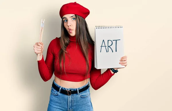 Young Brunette Teenager Holding Art Notebook Paintbrushes Making Fish Face — Stock Photo, Image