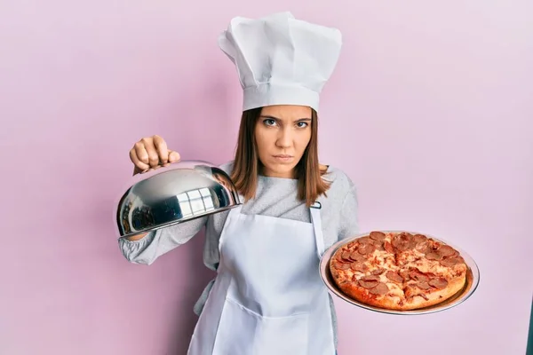 年轻而漂亮的女厨师拿着意大利披萨持怀疑和紧张的态度 皱着眉头因为问题而心烦意乱 消极的人 — 图库照片