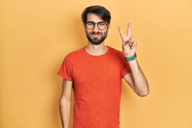 Gündelik kıyafetler giyen ve gözlüklü genç İspanyol adam iki numarayı gösterip işaret ederken kendinden emin ve mutlu bir şekilde gülümsüyor.. 