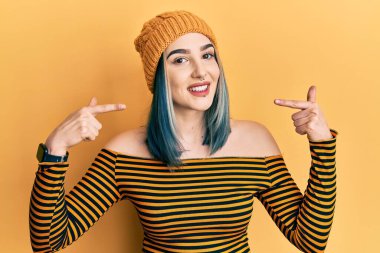 Yün şapka takan, neşeli gülümseyen, parmaklarıyla ve ağzıyla işaret eden genç, modern bir kız. diş sağlığı kavramı. 