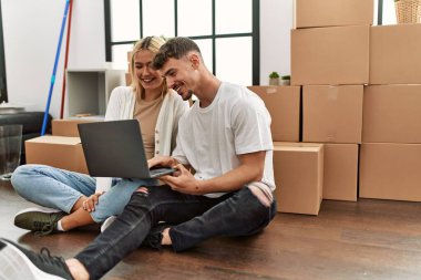 Genç beyaz çift yeni evlerinde dizüstü bilgisayar kullanarak mutlu bir şekilde gülümsüyor..