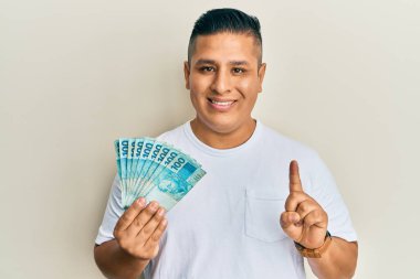 Elinde 100 Brezilyalı gerçek banknot tutan genç Latin bir adam bir fikir veya soru işareti ile gülümsüyor. 