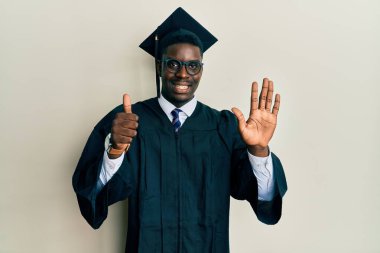Mezuniyet şapkası ve tören cübbesi takan yakışıklı siyah adam altı numaralı parmaklarıyla kendini beğenmiş ve mutlu gülümserken onu gösteriyor.. 
