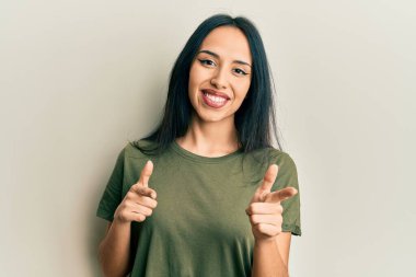 Günlük tişört giyen genç İspanyol kız mutlu ve komik yüzlü parmaklarını kameraya doğrultuyor. iyi enerji ve titreşimler. 