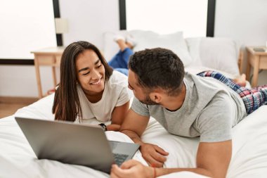 Yatakta dizüstü bilgisayar kullanarak mutlu gülümseyen genç Latin çift..