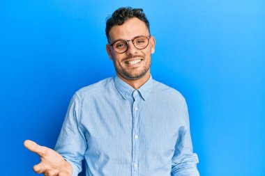 Gündelik kıyafetler ve gözlük takan genç İspanyol adam güler yüzlü ve kolları açık, dostça karşılama, olumlu ve kendinden emin selamlar. 