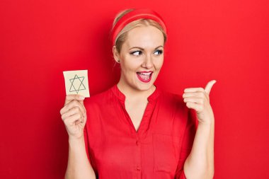 Sarışın genç bir kadın elinde Yahudi yıldızı hatırlatıcısıyla baş parmağını kaldırıyor ve ağzı açık gülümsüyor. 