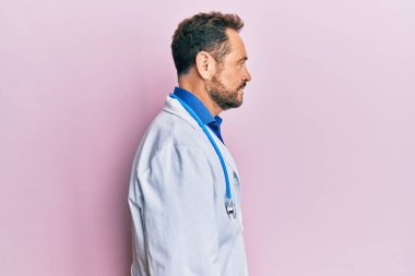 Doktor üniforması ve steteskop giyen orta yaşlı bir adam, rahat profil pozu verir, kendine güvenen bir gülümsemeyle doğal bir poz verir.. 