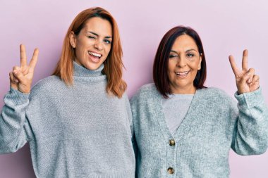 Latin anne ve kızı günlük kıyafetler giyip kameraya gülümsüyor ve parmaklarıyla zafer işareti yapıyor. İki numara.. 