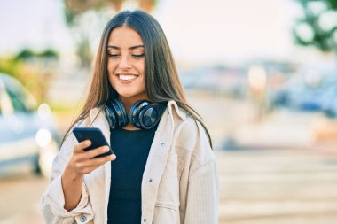 Genç İspanyol kız şehirde akıllı telefon ve kulaklık kullanarak mutlu bir şekilde gülümsüyor..
