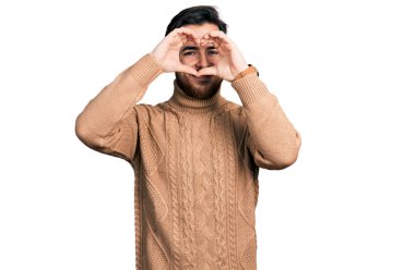 Gündelik giysiler giymiş, elleri ve parmaklarıyla kalp şeklinde gülümseyen genç İspanyol adam tabeladan bakıyor. 