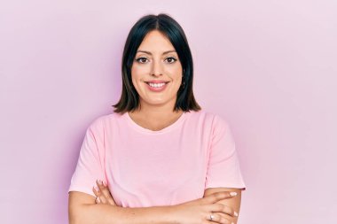 Gündelik pembe tişört giyen genç İspanyol kadın kollarını kavuşturup kameraya bakarken gülümsüyor. pozitif kişi. 
