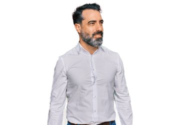 Orta yaşlı, sakallı, sıradan beyaz tişörtlü, yüzünde gülümseme ve doğal ifadeyle yan yana bakan bir adam. Kendinden emin gülen. 