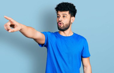 Sakallı, sıradan mavi tişörtlü genç bir Arap. Parmağı önde, ağzı açık, şaşırmış, ön tarafta bir şey var. 