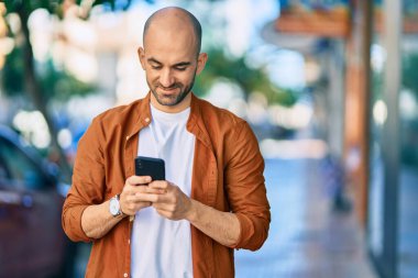Genç ve kel adam şehirde akıllı telefon kullanarak mutlu gülümsüyor..