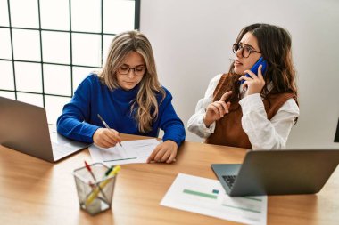 İki iş kadını akıllı telefonla konuşuyor ve ofiste dizüstü bilgisayar kullanıyor..
