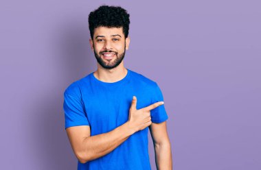 Sakallı, günlük mavi tişörtlü, neşeli, yüzünde bir gülümseme olan genç bir Arap adam, mutlu ve doğal bir ifadeyle elini ve parmağını kaldırıyor. 