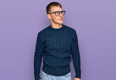 Sıradan kıyafetler ve gözlük takan genç sarışın adam yüzünde gülümsemeyle yan yana bakıyor, doğal bir ifade. Kendinden emin gülen. 