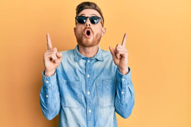 Şık güneş gözlüğü takan genç kızıl saçlı adam şaşırmış ve şaşırmış bir şekilde yukarı bakıyor ve parmaklarıyla işaret ediyor ve kollarını kaldırıyor.. 