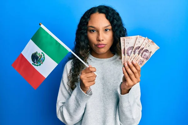 メキシコの国旗とメキシコペソ紙幣を持っている若いラテン語の女性は懐疑的で緊張しており 問題のために動揺していることを眉をひそめている 否定的な人 — ストック写真