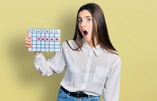 Young Brunette Teenager Holding Heart Calendar Scared Amazed Open Mouth — Stock Photo, Image