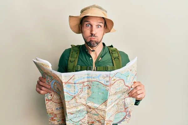 髭を生やしたハンサムな男が探検家の帽子をかぶって地図を持っていて面白い顔で頬をむいている 空気を吸い込み空気を吸い込み — ストック写真