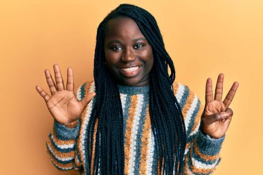 Saçları örgülü genç siyahi bir kadın, sıradan bir kazak giyiyor ve 8 numaralı parmaklarıyla işaret ederken, kendinden emin ve mutlu bir şekilde gülümsüyor.. 