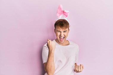 Paskalya tavşanı kulakları takan genç beyaz adam kolları havada ve gözleri kapalı olarak başarıyı kutluyor ve şaşırıyor. 