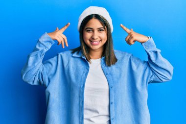 Hoş yün şapkalı genç Latin kadın iki eliyle başını işaret ederek gülümsüyor, harika bir fikir ya da düşünce, iyi bir hafıza 