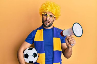 Genç İspanyol futbolcu futbol taraftarı elinde futbol topu tutarken megafondan bihaber ve kafası karışık bir ifade kullanıyor. şüphe kavramı. 