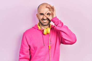 Spor kıyafetleri giyen ve kulaklık takan genç kel bir adam. El hareketleriyle, gülümseyerek, gözleriyle mutlu yüzüyle parmaklarının arasından bakıyor.. 