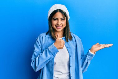 Güzel yün şapkalı genç Latin kadın avuçlarını gösteriyor ve baş parmaklarıyla iyi bir jest yapıyor, mutlu ve neşeli gülümsüyor. 