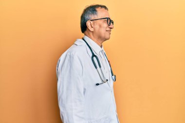 Orta yaşlı Hintli bir adam doktor önlüğü ve steteskop takıyor, rahat profil pozu veriyor, kendine güvenen bir gülümsemesi var.. 