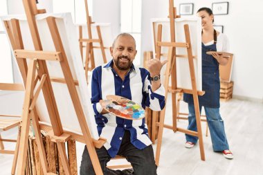 Sanat stüdyosundaki kıdemli sanatçı arka tarafı işaret ediyor. Elleri havada, kendinden emin gülümsüyor. 