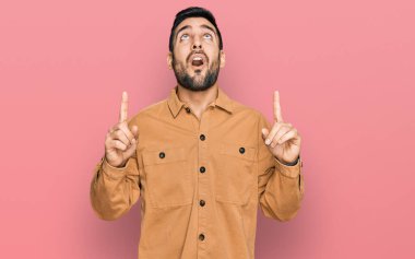 Gündelik giysiler giyen genç İspanyol adam şaşırmış ve şaşırmış bir şekilde yukarı bakıyor ve parmaklarıyla işaret ediyor ve kollarını kaldırıyor.. 